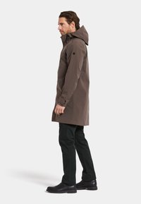 Brun vandtæt parka med hætte, sidelommer og en længere skæring, parret med mørkegrønne bukser og sorte støvler. Glat tekstur.