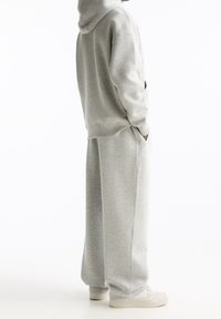 Person iført en lys grå hættebluse og matchende brede sweatpants med hvide sneakers, stående sidelæns med hænderne i lommerne.