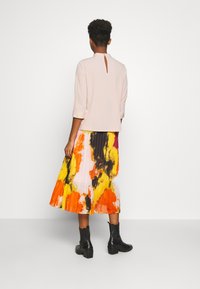 Haut rose clair avec un détail en forme de goutte dans le dos ; associé à une jupe plissée colorée arborant des motifs abstraits en orange, jaune et noir. Bottines noires.