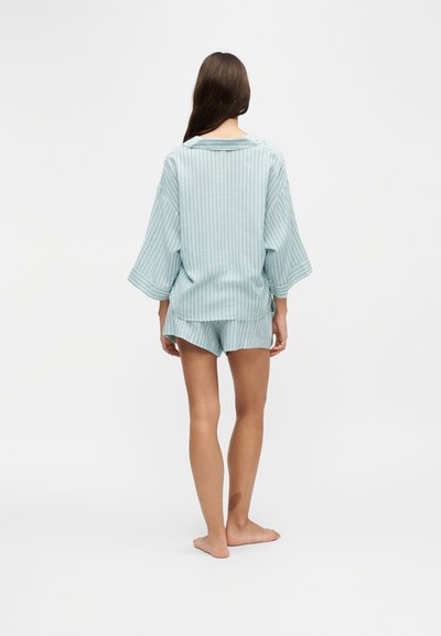 Femme debout, pieds nus, de dos, portant un haut à manches longues rayé bleu clair et un short assorti sur fond blanc.