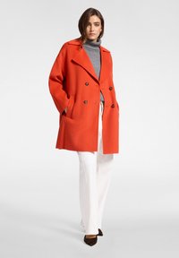 Cappotto doppiopetto in lana arancione con grandi revers, tasche e bottoni marroni, abbinato a un dolcevita grigio e pantaloni bianchi.