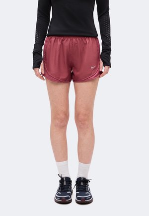 Osoba nosząca bordowe szorty do biegania marki Nike, czarną bluzę z długim rękawem z otworami na kciuki, białe skarpetki i czarne buty do biegania, stojąca prosto.