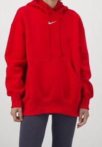 Röd Nike-hoodie i mjukt tyg, med en ficka fram, dragsnören och en liten vit logotyp på bröstet. Löst snitt.