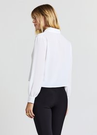 Weiße, kurze Bluse mit langen, voluminösen Ärmeln und einem Kragen; glatter, leichter Stoff; Rückansicht vor neutralem Hintergrund.