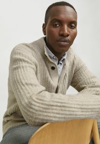 Beige gerippter Strickcardigan mit strukturierter Oberfläche und Knopfverschluss, über einem blau-weißen gestreiften Hemd getragen.