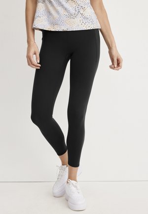 Woman wearing black leggings, white platform sneakers, and a patterned top standing against a white background.