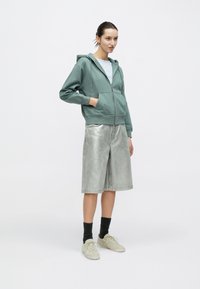 Πράσινο hoodie με φερμουάρ, ανοιχτό μπλε μπλουζάκι, ασημί τζιν σορτς και μπεζ αθλητικά παπούτσια. Το μοντέλο στέκεται μπροστά σε ένα απλό λευκό φόντο.