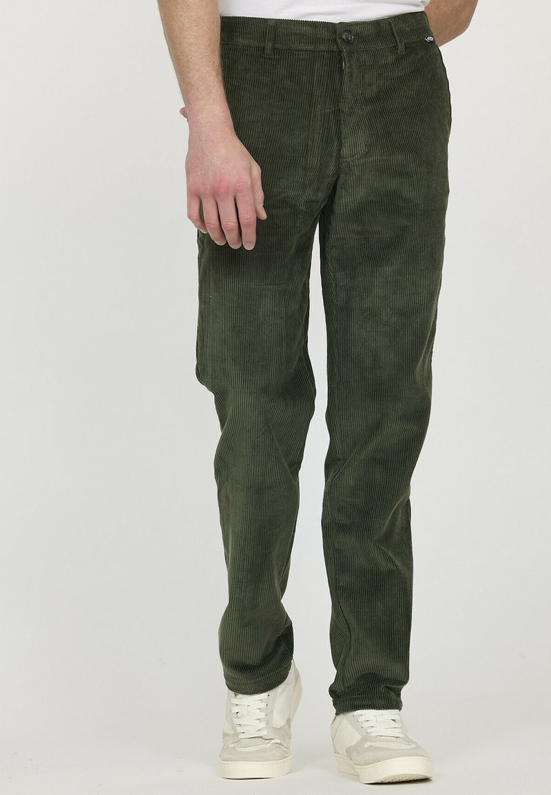 Pantalons en velours côtelé vert foncé avec des côtes verticales, des poches avant et une fermeture à bouton. Portés avec des baskets blanches.