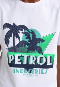 Camiseta blanca con un diseño gráfico que presenta palmeras de color azul oscuro, formas geométricas verdes y el texto "PETROL INDUSTRIES" en letras gruesas.
