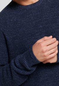 Marineblauer Strickpullover mit geripptem Muster, rundem Ausschnitt und verlängerten Bündchen. Der Stoff hat ein leicht flauschiges Aussehen.