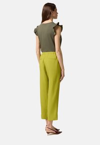 Haut vert olive avec des manches volantes, associé à un pantalon vert clair avec une taille ajustée et une poche arrière, complété par des talons bruns.
