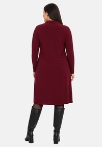 Robe bordeaux à manches longues avec un design fluide, fabriquée en tissu doux. Assortie à des bottes noires brillantes jusqu'aux genoux avec des talons épais.