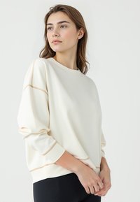 Sweatshirt crème avec un col rond, des épaules tombantes et des coutures décoratives couleur tan. Tissu doux avec une coupe décontractée.