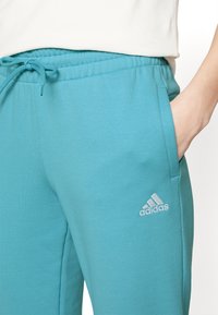 Teal sweatpants made of soft fabric with an elastic waistband, drawstring, and side pockets. Features a white Adidas logo on the left leg.