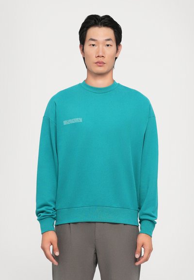 Tyrkysový crewneck sveter s rebrovanými manžetami a lemom; na prednej strane má malý biely textový detail. Hladká, jednoliata textúra.