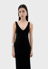 Zwart geribbeld maxi-jurk met een diepe V-hals, mouwloos ontwerp en een aansluitende silhouet. Model draagt een zilveren manchette.