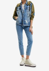 Chaqueta de denim con mangas verde oliva, detalles desgastados y acentos de logo. Combinada con jeans azul claro y zapatillas altas de patrón beige.