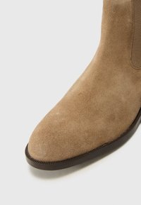 Bottine en cuir suédé marron avec un bout arrondi et des panneaux latéraux élastiques. Texture lisse avec une semelle foncée contrastante.