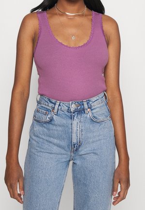 Femme portant un haut sans manches violet côtelé avec bordure en dentelle et un jean taille haute bleu clair, accessoirisée avec des colliers dorés superposés.