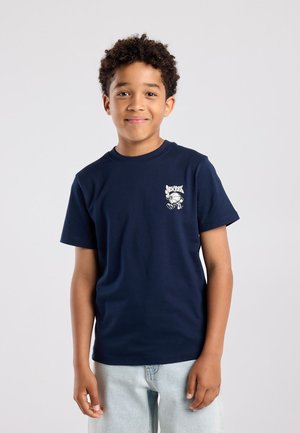 Jongen met krullend haar lacht, draagt een donkerblauw T-shirt met een kleine New York-afbeelding en lichtblauwe spijkerbroek, staand tegen een effen achtergrond.