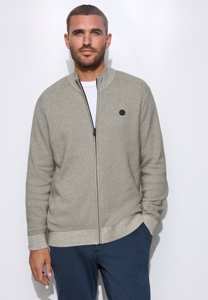 Homme aux cheveux courts et à la barbe, portant un pull gris clair à fermeture éclair sur une chemise blanche et un pantalon bleu marine, posant devant un fond blanc uni.