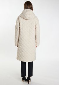 Beige kviltet lang jakke med hætte, med et diamantmønster. Båret over sorte bukser og højhælede sko. Glat tekstur.