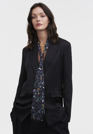 Donna con capelli scuri di lunghezza spalla, che indossa un blazer nero e una sciarpa di seta nera a fantasia floreale annodata in modo sciolto attorno al collo.