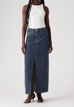 Kobieta w białym, bezrękawnikowym topie oraz granatowej, długiej plisowanej spódnicy maxi z pionowymi paskami i rozcięciem z przodu, nosząca czarne sandały na obcasie.