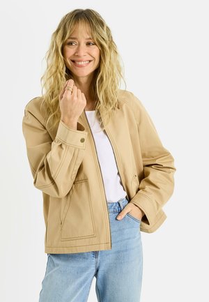 Mujer sonriente con cabello rubio ondulado que lleva una chaqueta beige, camisa blanca, vaqueros azules y anillos en la mano izquierda sobre un fondo blanco liso.
