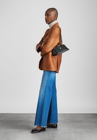 Smeđi kožni blejzer preko bijelog turtlenecka, uparen sa širokim plavim trapericama i crnim mokasinkama. Crna torba s zlatnim detaljima.