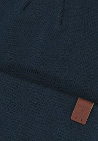 Granatowa czapka z dzianiny, z teksturą prążkowaną. Posiada małą brązową skórzaną metkę z wytłoczoną nazwą marki.