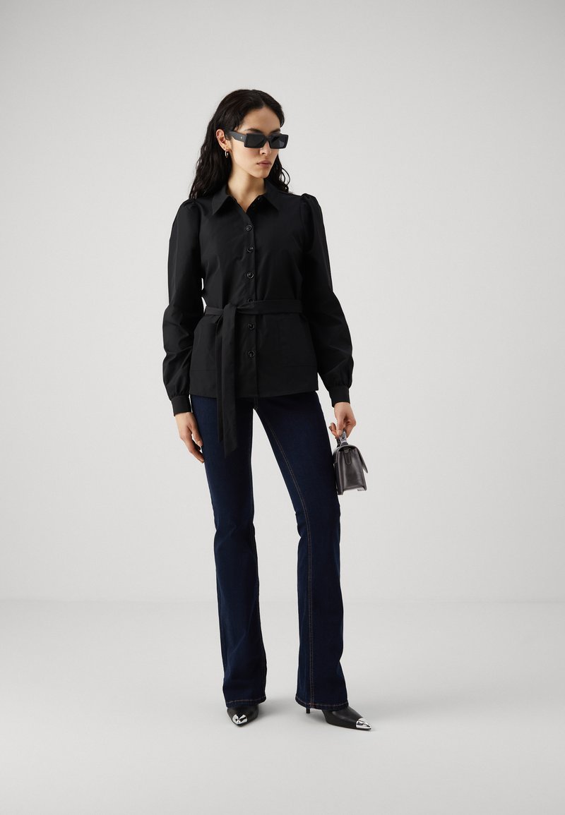 Schwarze Bluse mit Knopfverschluss und gebundenem Bund, langen puffärmeligen Ärmeln, kombiniert mit ausgestellten Jeans und spitzen Absatzschuhen. Die Accessoires umfassen dunkle Sonnenbrillen.