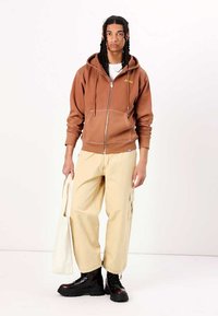 Brown zip-up hoodie with front pockets, paired with loose-fitting beige cargo pants and black lace-up boots. Holding a white tote bag.