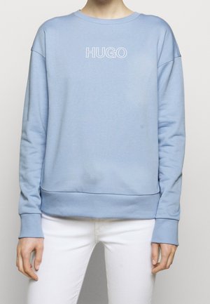 Person wearing a light blue sweatshirt with "HUGO" text on chest and white pants, standing against a plain light background.