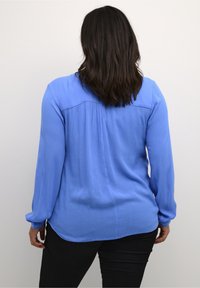 Blusa de manga larga azul claro con una tela texturizada, detalle de pliegues en la parte posterior y dobladillo curvado, combinada con pantalones ajustados negros.