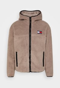 Giacca in pile beige con cappuccio, zip frontale completa e dettagli neri. Presenta una toppa con logo sul petto e due tasche laterali con zip.