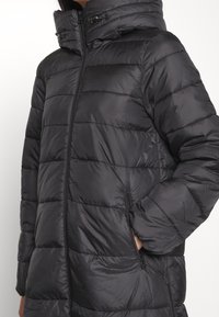 Černá prošívaná bunda s vysokým límcem a kapucí. Má prošívané části, zapínání na zip vpředu a boční kapsy. Měkký, lehký materiál.