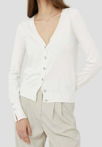 Vit kofta i mjukt stickat material. Har djup V-ringning, långa ärmar och silverknappar. Diskret logotyp nära fållen.