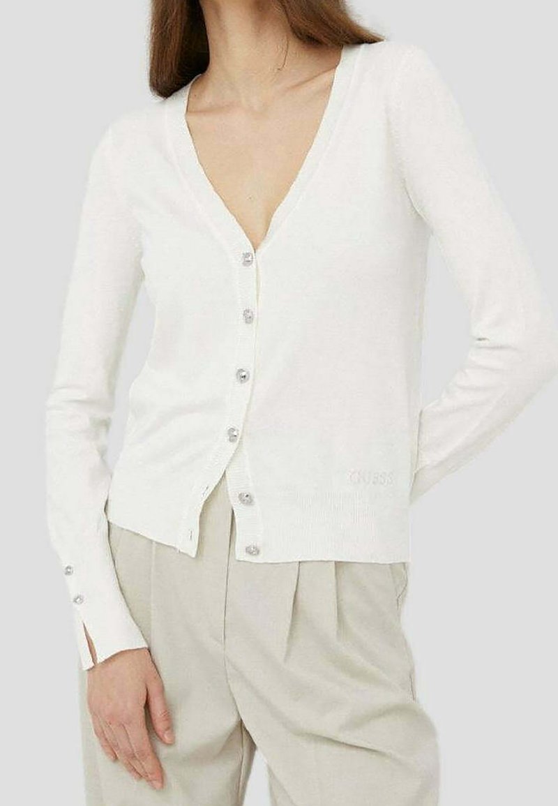 Vit kofta i mjukt stickat material. Har djup V-ringning, långa ärmar och silverknappar. Diskret logotyp nära fållen.