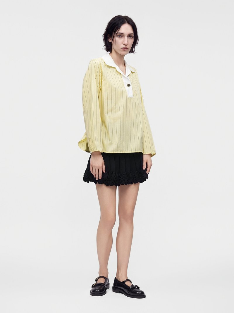 Femme aux cheveux courts et foncés portant une chemise jaune ample à manches longues avec un col blanc, une mini-jupe noire à volants et des chaussures noires, debout.