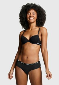 Lächelnde Frau mit lockigem Haar, die einen schwarzen Puma-BH und passende schwarze Puma-Unterwäsche vor einem schlichten hellen Hintergrund trägt.