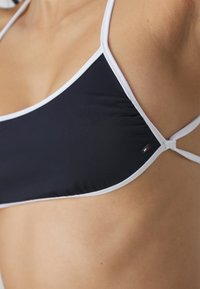 Námornícky bikiny top s bielym lemem, s nastaviteľnými popruhmi a akcentom loga. Hladká tkanina s priliehavým strihom.