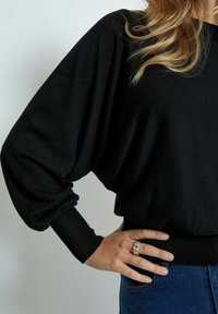 Musta ylipitkä neule, jossa on väljät hihat ja resorikainalot, valmistettu pehmeästä kankaasta. Yhdistetty sinisiin farkkuihin.