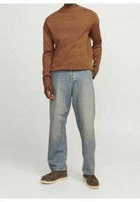 Brauner Strickpullover mit rundem Ausschnitt, kombiniert mit hellblauen, locker sitzenden Jeans. Das Modell trägt dunkle Schuhe mit hellen Sohlen.