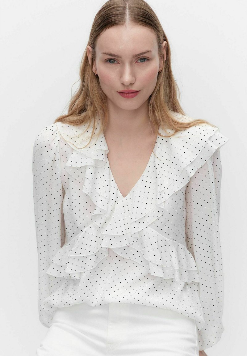 Jonge vrouw met lang blond haar, gekleed in een wit bloesje met zwarte stippen en ruches, poserend tegen een effen achtergrond.