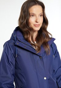 Giacca impermeabile blu navy con cappuccio, chiusura con zip e bottoni, cordini e tasche laterali. Tessuto liscio e design aderente.