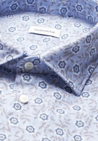 Chemise bleu clair avec petit motif floral bleu et gris, boutons blancs et étiquette « seidensticker » à l’intérieur du col.