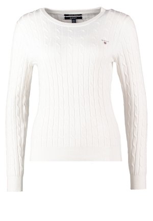 Femme marchant portant un pull en maille torsadée blanche, un jean skinny clair déchiré, des chaussettes noires et des baskets bleu marine sur un fond uni.
