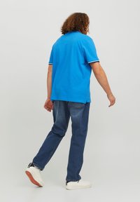 Person with curly hair wearing a blue polo shirt, blue jeans, white sneakers, walking against a plain light gray background.