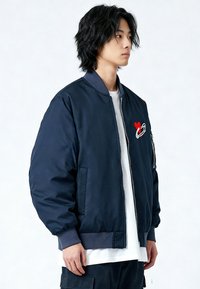 Giacca bomber navy con zip frontale, polsini a costine e dettaglio ricamato a forma di cuore e spilla da balia sul petto. Tessuto liscio e imbottito.